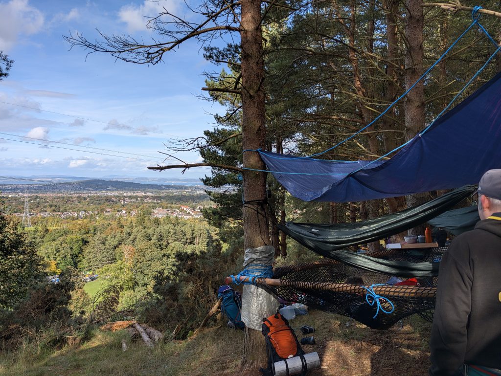 A SkyCamp base lashed between pine trees, with a blue tarp, hammocks and a cargo net, looking out over Edinburgh